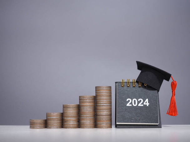 Μελέτη στόχους, 2024 Ημερολόγιο γραφείου με καπέλο αποφοίτησης και στοίβα των κερμάτων. Η έννοια της εξοικονόμησης χρημάτων για την εκπαίδευση, φοιτητικό δάνειο, υποτροφία, δίδακτρα το νέο έτος 2024 - Φωτογραφία, εικόνα