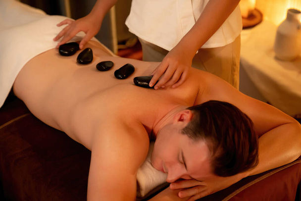 Hot stone massage in spa salon in luxe resort met warme kaars licht, gelukzalige man klant genieten spa basalt steen massage glijden over het lichaam met rustgevende warmte. Stil maar. - Foto, afbeelding