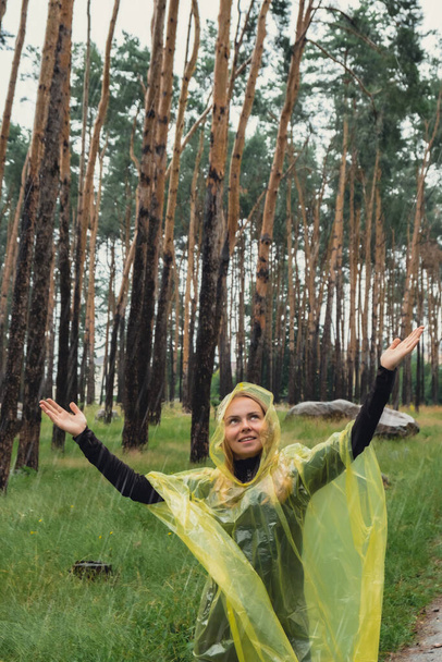 Mujer sonriente en impermeable amarillo caminando en el bosque de otoño disfrutando del clima lluvioso al aire libre. Turista femenina descubre parque en temporada de lluvias. Cercanía a la naturaleza. Satisfacción de curación mental aire limpio  - Foto, imagen