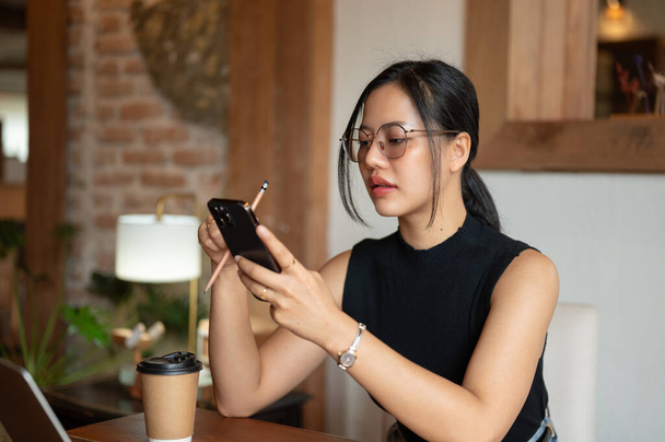 Een mooie duizendjarige Aziatische vrouw gebruikt haar smartphone, reageert op berichten, of scrollt op sociale media terwijl ze in een mooie coffeeshop zit. - Foto, afbeelding
