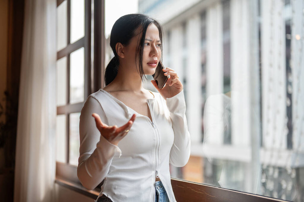 Una donna asiatica arrabbiata e insoddisfatta sta parlando al telefono mentre sta vicino alla finestra, avendo una discussione al telefono con qualcuno.. - Foto, immagini