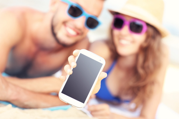 Happy couple showing smartphone at the beach - 写真・画像