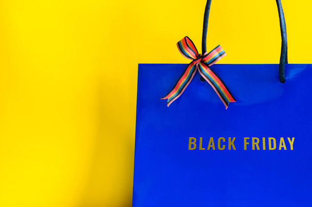 ブラックフライデーショッピングコンセプトの黄色の背景にリボンとブラックフライデーの言葉のブルーショッピングバッグ. - 写真・画像