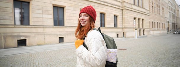 Tourism and travelling. Young redhead woman smiling, tourist walking with backpack around city centre, sightseeing, holds backpack with paper map. - Foto, Imagem