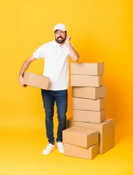 Full-length tiro de homem de entrega entre caixas sobre fundo amarelo isolado frustrado e cobrindo orelhas - Foto, Imagem