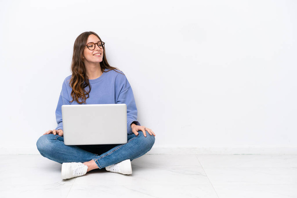 Fiatal kaukázusi nő egy laptop ül a padlón elszigetelt fehér háttér néz oldalra, és mosolyog - Fotó, kép