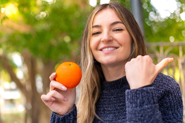 Young pretty Romanian woman holding an orange at outdoors pointing to the side to present a product - Photo, Image