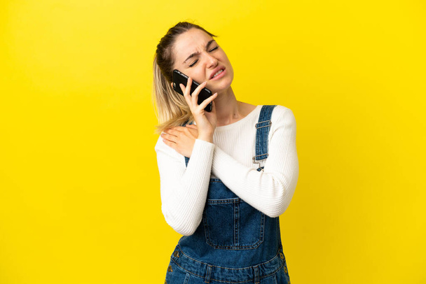 Junge Frau benutzt Handy über isoliertem gelben Hintergrund und leidet unter Schmerzen in der Schulter, weil sie sich bemüht hat - Foto, Bild
