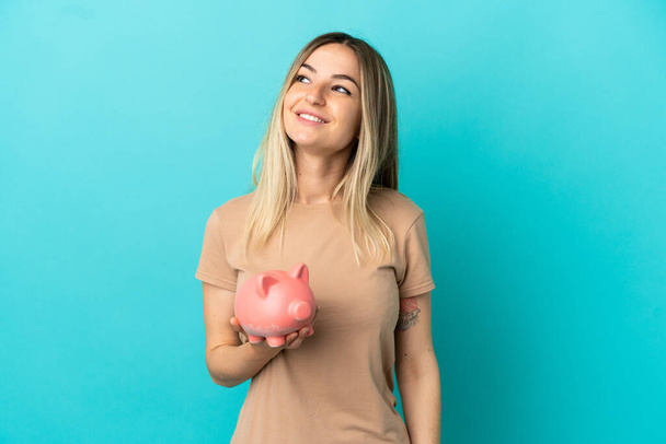 Young woman holding a piggybank over isolated blue background thinking an idea while looking up - Photo, Image