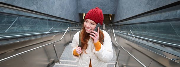 Portrait of young redhead woman walks around city, goes up stairs with mobile phone, talks on smartphone and smiles. - Foto, afbeelding