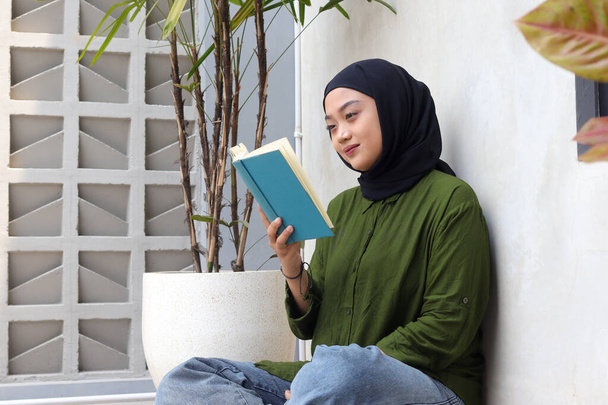 Retrato de la atractiva mujer asiática hijab leyendo libro en balcón. Chica musulmana disfrutando del tiempo libre. Concepto de alfabetización y conocimiento - Foto, imagen