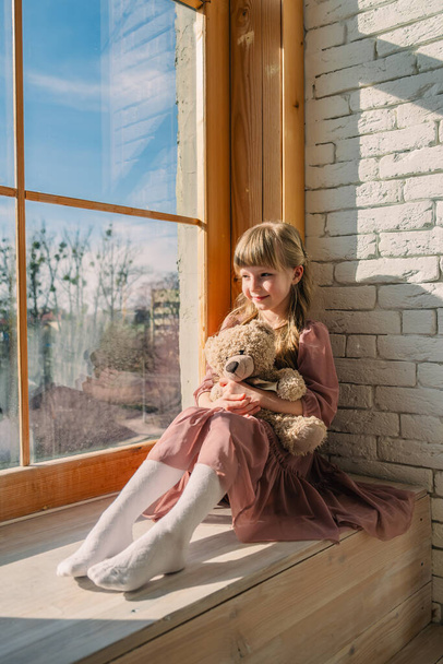 piccola ragazza carina in abito vicino alla finestra con orso giocattolo - Foto, immagini