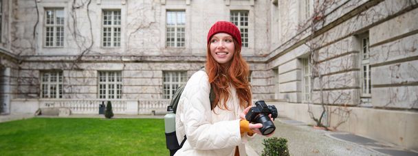 Happy redhead girl tourist, takes photos, photographer with professional camera walks around city and captures beautiful pictures. - Foto, Imagem