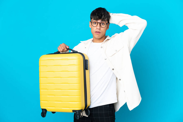 Mladý muž přes izolované modré pozadí v dovolené s cestovním kufrem a překvapen - Fotografie, Obrázek