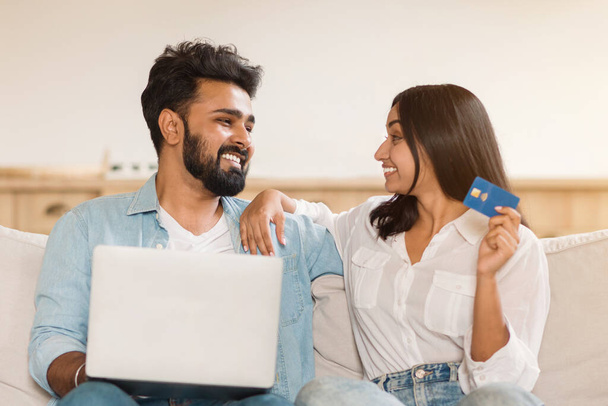 Молодая индийская пара наслаждается временем вместе, мужчина с помощью ноутбука, женщина держит кредитную карту, глядя друг на друга и улыбаясь, отдыхая на диване дома - Фото, изображение