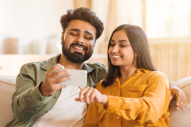 Genç Hintli çift evde rahat bir şekilde oturup selfie çekerken keyifli anlarını paylaşıyorlar. Her ikisi de samimi gülücükler sergiliyor, samimi ve zevkli paylaşılan bir deneyim yakalıyorlar. - Fotoğraf, Görsel