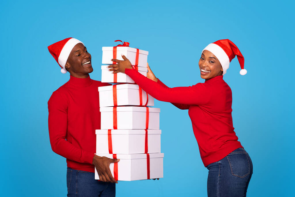 Jeune couple afro-américain manipulant des piles de boîtes-cadeaux avec des rubans rouges, fond bleu. Conjoints appréciant l'excitation des échanges présents de Noël et des célébrations festives - Photo, image