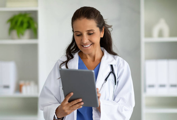 Modern technológiák az egészségügyben. Mosolygó érett nő orvos digitális tabletta pózol az irodájában, másolás helyet. Happy Lady terapeuta gazdaság digitális pad, klinika belső - Fotó, kép