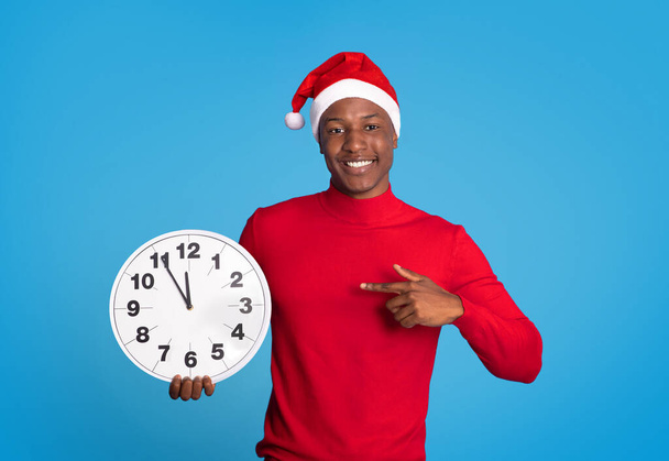 Black Man In Festive Santa Hat Pointant doigt à l'horloge debout sur fond bleu en studio, compte à rebours minutes de l'année à venir. L'excitation de Noël et des festivités du Nouvel An - Photo, image
