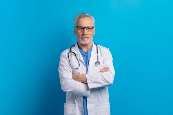 Portret van een zelfverzekerde, knappe, grijsharige, oudere arts in medisch uniform met stethoscoop en bril, armen gekruist op de borst, geïsoleerd op een blauwe studioachtergrond - Foto, afbeelding
