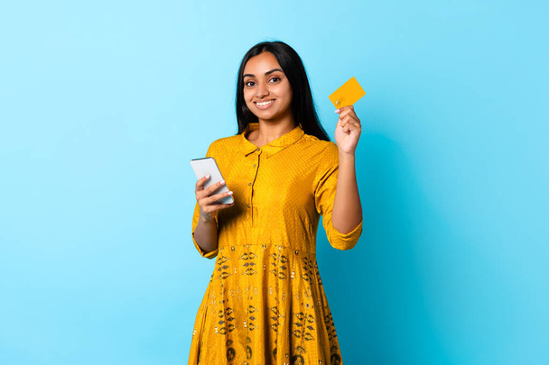Jeune femme hindoue souriante en robe jaune à l'aide d'une carte de débit et d'un smartphone pour faire du shopping en ligne sur fond de studio bleu. Faire place au commerce électronique moderne et aux applications bancaires - Photo, image