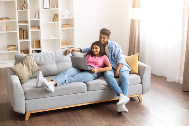 Young Indian couple enjoys leisurely day on their cozy sofa, engaging with laptop together in warmly lit, book-filled living room, full length, free space - Photo, Image