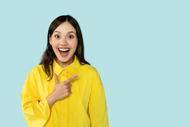Emocionální veselá hezká mladá žena ukazující na kopírovací prostor a usmívající se. Happy dáma ukazuje skvělou nabídku, řešení, na sobě žluté tričko, izolované na modrém pozadí studia - Fotografie, Obrázek