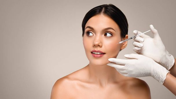Cosmetologist professional in white gloves make injection to millennial caucasian lady, look at copy space, isolated on gray studio background, panorama. Beauty esthetic care, cosmetic procedures - Photo, Image