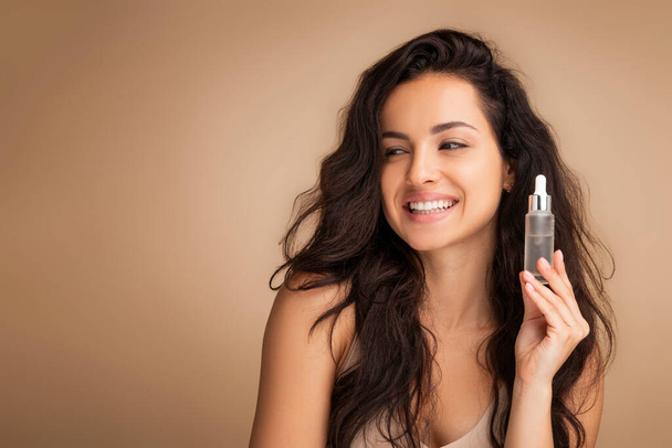 Anti-Age Cosmetics. Attractive Millennial Woman Holding Bottle With Face Serum, Happy Smiling Indian Lady Moisturizing Skin While Standing Over Light Background. Looking At Copy Space - Fotografie, Obrázek