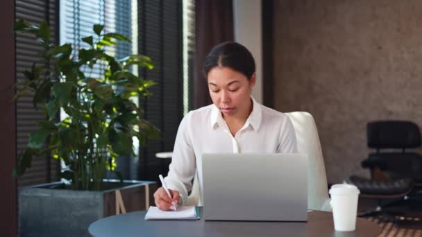 Συγκεντρωμένη πολυπολιτισμική γυναίκα κάθεται σε στρογγυλό τραπέζι μπροστά από το σύγχρονο φορητό υπολογιστή, κοιτάζοντας την οθόνη και αντιγραφή πληροφοριών από το διαδίκτυο στο φορητό υπολογιστή. Έννοια της τεχνολογίας και της εργασίας γραφείου. - Πλάνα, βίντεο