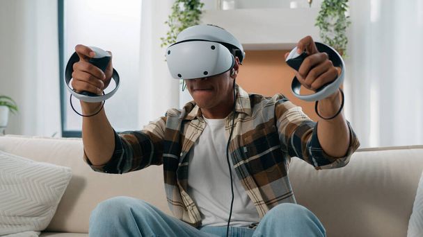 Αφρο-Αμερικανός άνθρωπος που διασκεδάζει στο σπίτι καναπέ με εικονική πραγματικότητα παιχνίδι αγώνα τύπος παιχνίδι 3D κυβερνο-gaming καταπολέμηση με χειριστήρια άνθρωπος που παίζει σε VR γυαλιά κράνος χρησιμοποιώντας joysticks metaverse κόσμο - Φωτογραφία, εικόνα