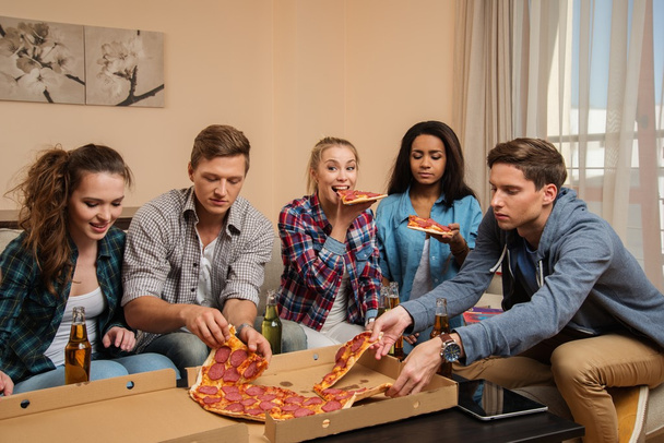 Gruppe junger multiethnischer Freunde feiert mit Pizza und Getränkeflaschen im heimischen Interieur - Foto, Bild