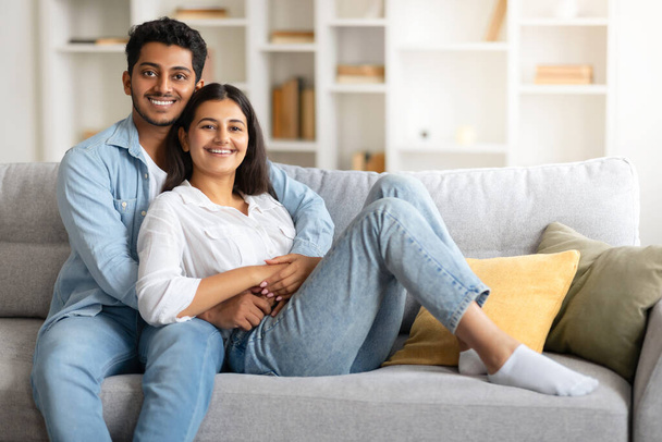 Onnellinen rakastava intialainen pari kotona, aviomies syleilee vaimoaan kodikas sohva, sekä iloisesti hymyillen kameralle, kapseloi kodikas ja rauhassa ilmapiiri, vapaa tila - Valokuva, kuva