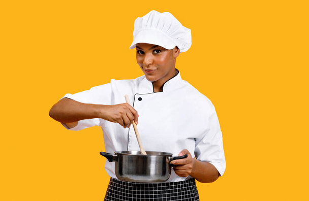 Концепція приготування їжі. Усміхнена чорна шеф-кухарка, що перемішує їжу в каструлі, стоячи ізольовано над жовтим тлом, молода афроамериканська кухарка леді в уніформі, використовуючи дерев'яний шпатель, готуючи їжу - Фото, зображення