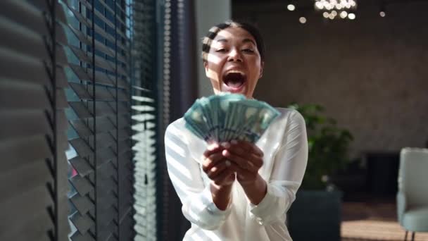 Mujer multiracial con sonrisa radiante mirando a la cámara y sosteniendo ventilador de billetes de dólar. Exitosa dama de negocios regocijando los ingresos del dinero mientras está de pie cerca de la ventana de la oficina con persianas medio cerradas. - Metraje, vídeo