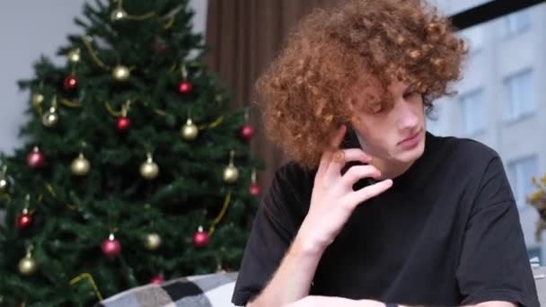 Młody mężczyzna rozmawia przez telefon przy kolorowym drzewie zimowym w pokoju. Świąteczne pozdrowienia i prezenty. - Materiał filmowy, wideo