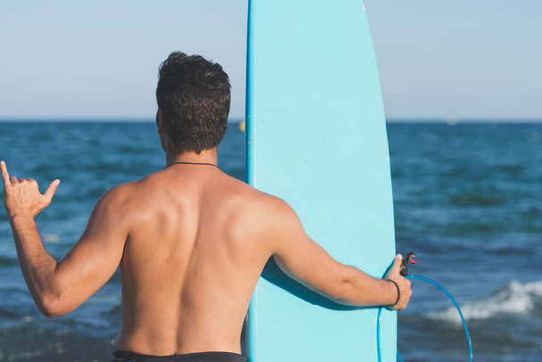 Neoprene mayo giymiş yakışıklı ve çekici bir sörfçü elinde sörf tahtası, yaz sporu ve tatillerin tadını çıkardıktan sonra havalı poz veriyor. - Fotoğraf, Görsel