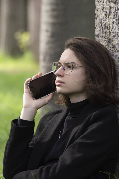 Student v brýlích, obchodní bundě a džínách sedí na trávě v parku, s telefonem u ucha, zamyšleně a upřeně hledí do dálky na pozadí přírody v parku. - Fotografie, Obrázek