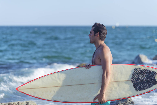 Un hombre con el torso desnudo parado en la playa sosteniendo una tabla de surf - Foto, imagen