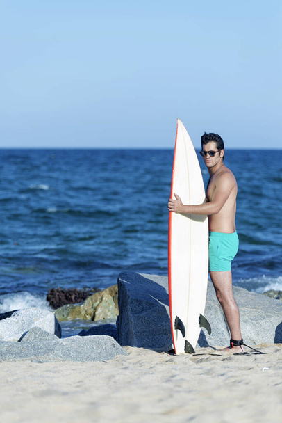 hombre surfista guapo y atractivo en traje de baño de neopreno sosteniendo tabla de surf posando fresco después de surfear disfrutando del deporte acuático de verano y vacaciones - Foto, Imagen