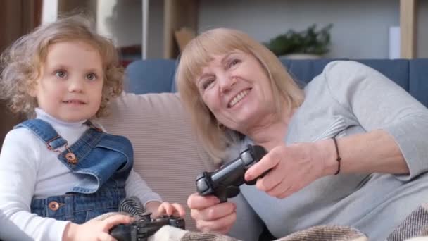 Щаслива сучасна бабуся середнього віку, що сидить на дивані з маленькою онучкою, грає в комп'ютерну гру разом, щаслива старша бабуся розважається з маленьким онуком. - Кадри, відео