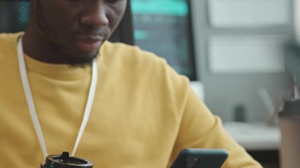 Kallista kuvaa nuoresta afroamerikkalaisesta nörtistä älypuhelimella ja kahvikupilla, joka istuu toimistossa työpisteessä - Materiaali, video