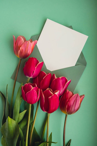 Empty white envelope copy space for your text or design with beautiful pink tulip bouquet on green background. Template mock up for holiday spring greeting card. Top view Flat lay blank paper Feminine - Photo, Image