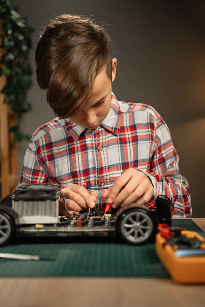 Teen boy sitting at table and soldering remote controlled car. Caucasian child fixing favorite toy by himself at home. Copy space - Photo, Image
