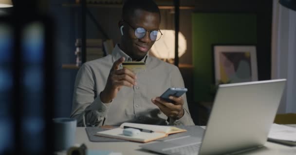 African American businessman using smartphone and credit card making payment in dark office at night. Banking and finance concept. - Felvétel, videó