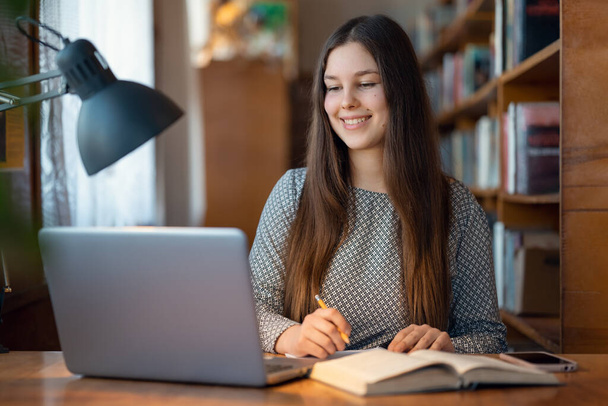 Χαριτωμένη νεαρή μαθήτρια κάθεται σε ένα τραπέζι με φορητό υπολογιστή μπροστά της, γράφοντας κάτι στο σημειωματάριο. Έννοια της εκπαίδευσης, σπουδές σε βιβλιοθήκη, σύγχρονοι φοιτητές - Φωτογραφία, εικόνα