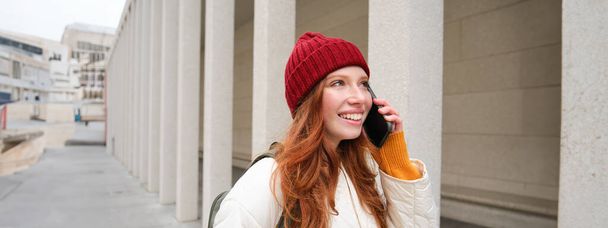 Mobile broadband and people. Smiling young redhead woman walks in town and talks on mobile phone, calling friend on smartphone, using internet to make a call abroad. - Photo, image