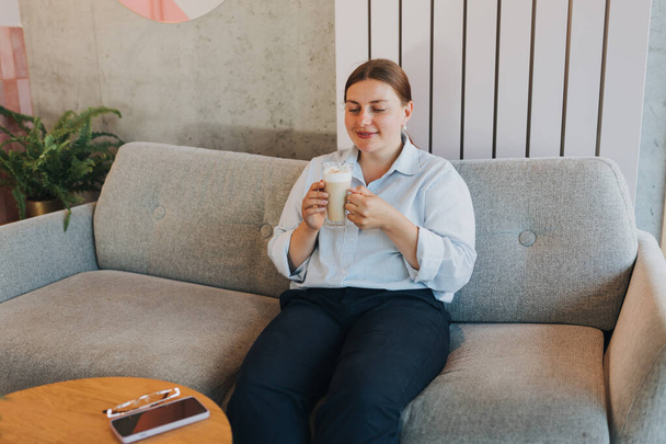 Onnellinen liikenainen, joka pitelee kuppia sohvalla kotona. 30s nainen lasit kahvilassa kahvin juominen - Valokuva, kuva