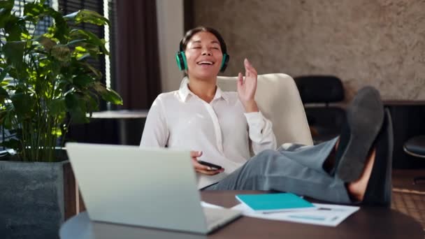 Χαρούμενη πολυφυλετική γυναίκα που κάθεται στο χώρο εργασίας με τα πόδια στο γραφείο και διασκεδάζει ακούγοντας μουσική σε ασύρματα ακουστικά. Ευτυχισμένος υπάλληλος γραφείου ψάχνει το αγαπημένο τραγούδι στο σύγχρονο smartphone στα χέρια. - Πλάνα, βίντεο