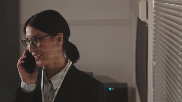 Cintura para cima de uma jovem trabalhadora de segurança cibernética olhando através de persianas em seu escritório enquanto faz uma chamada telefônica privada no local de trabalho tarde da noite - Filmagem, Vídeo